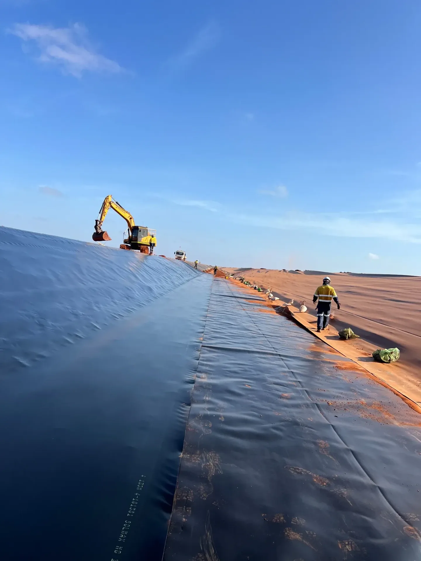 HDPE geomembrane liner installed down a long batter slope with an excavator and crew on site